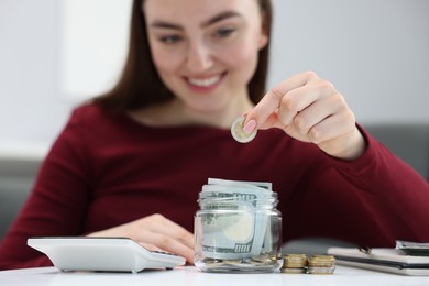 Photo of Woman putting coin into glass jar at table indoors, selective focus