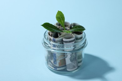 Photo of Glass jar with dollars and sprout on light blue background, closeup