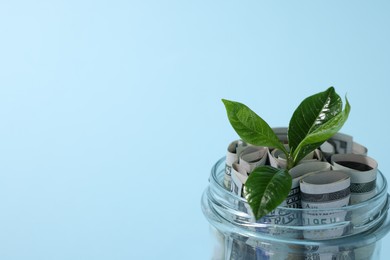 Photo of Glass jar with dollars and sprout on light blue background, closeup. Space for text