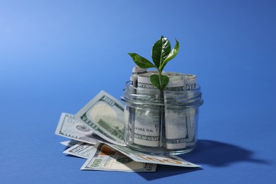 Photo of Glass jar with dollars and sprout on blue background, closeup