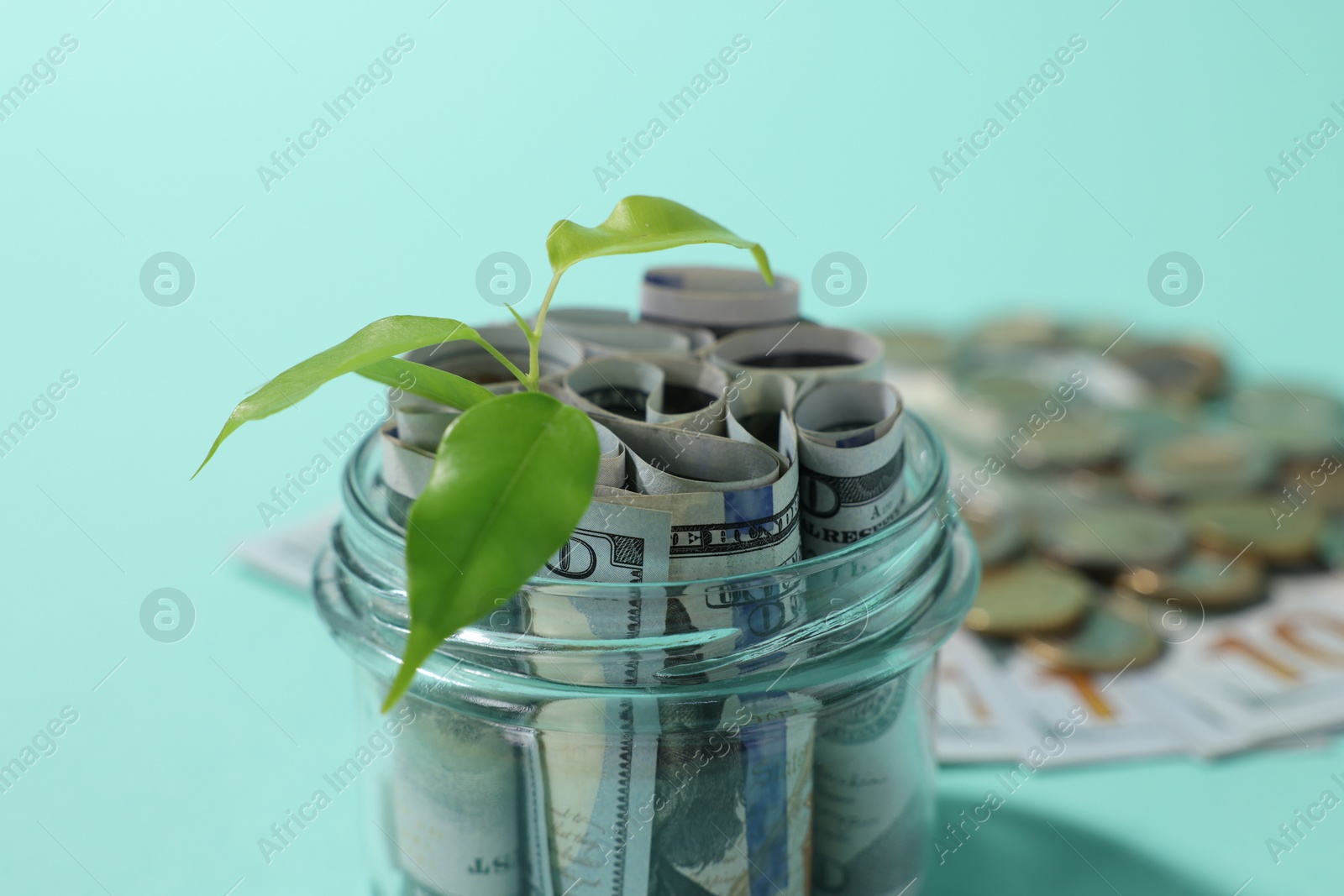 Photo of Glass jar with dollars, sprout and coins on light blue background, closeup