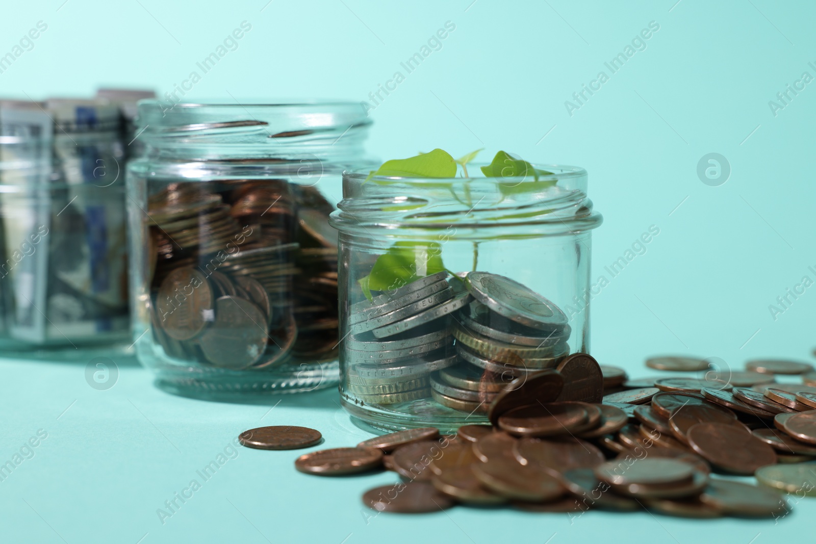 Photo of Glass jars with coins, sprout and dollars on light blue background, closeup