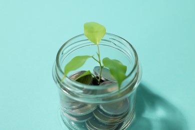 Photo of Glass jar with coins and sprout on light blue background, closeup