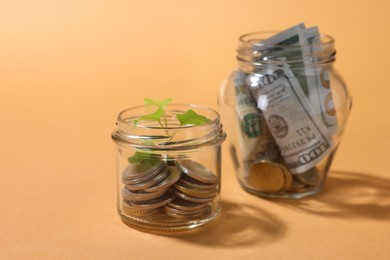 Photo of Glass jars with coins, sprout and dollars on pale orange background, closeup