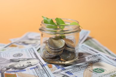Photo of Glass jar with coins, sprout and dollars on pale orange background, closeup