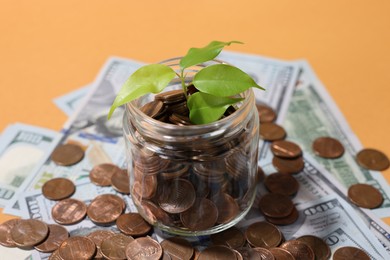 Photo of Glass jar with coins, sprout and dollars on pale orange background, closeup