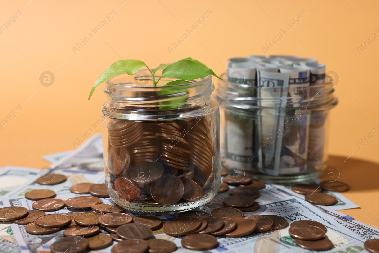 Photo of Glass jars with coins, sprout and dollars on pale orange background, closeup