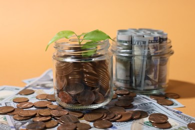Photo of Glass jars with coins, sprout and dollars on pale orange background, closeup