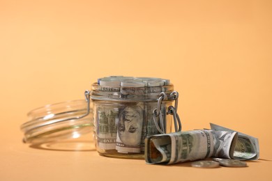 Photo of Glass jar with dollars and coins on pale orange background, closeup