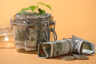 Photo of Glass jar with dollars, coins and sprout on pale orange background, closeup