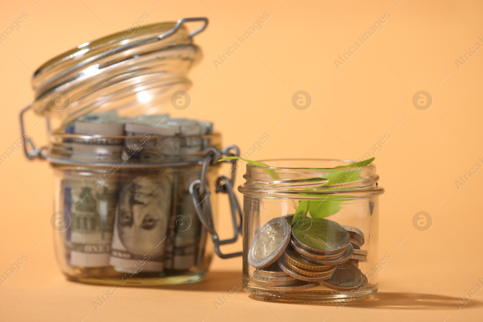 Photo of Glass jars with dollars, coins and sprout on pale orange background, closeup