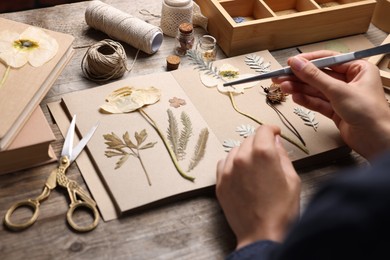 Photo of Woman putting dry flowers into notebook at wooden table, closeup