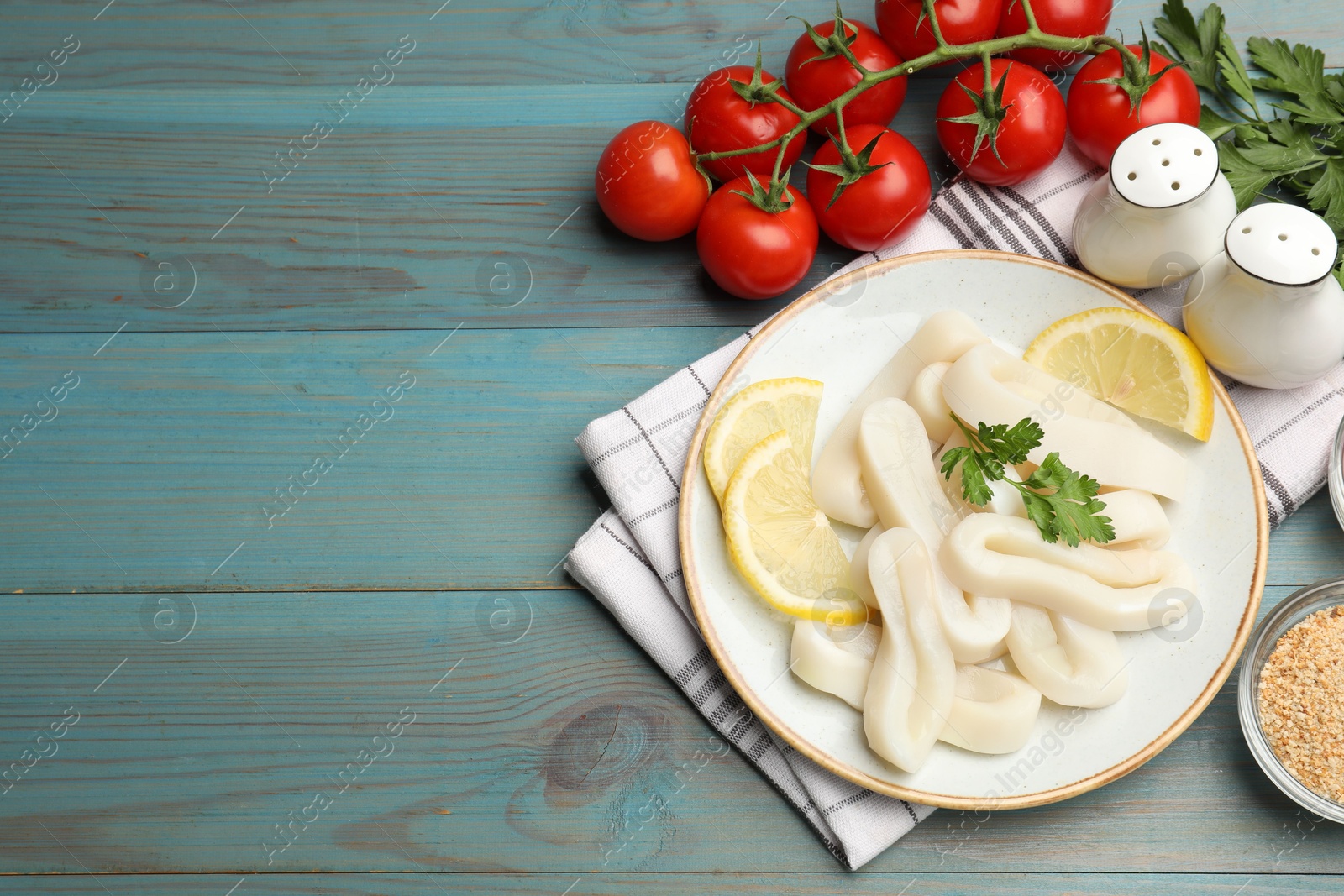 Photo of Raw squid rings, parsley, lemon, spices and tomatoes on light blue wooden table, flat lay. Space for text