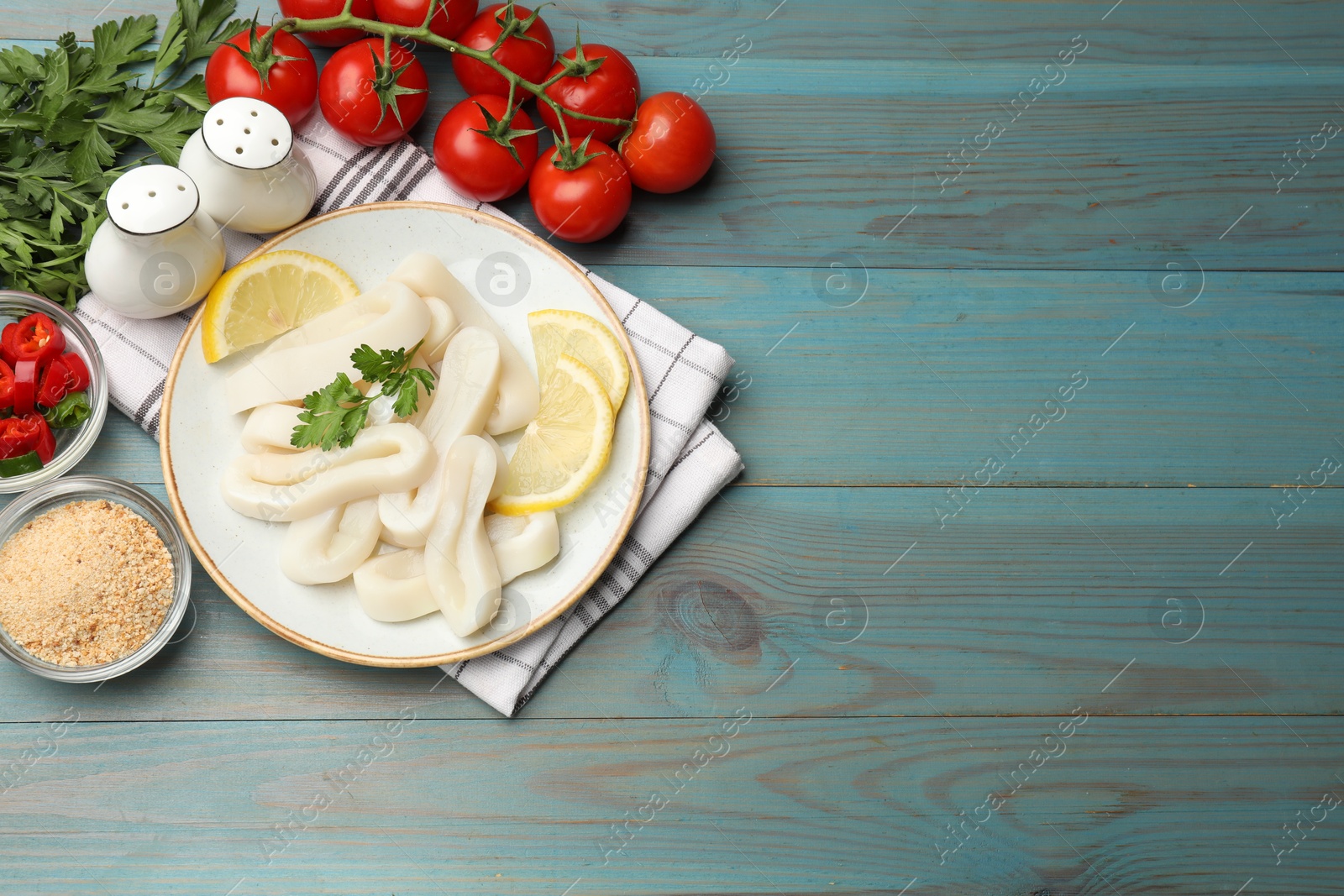 Photo of Raw squid rings, parsley, lemon, spices and tomatoes on light blue wooden table, flat lay. Space for text