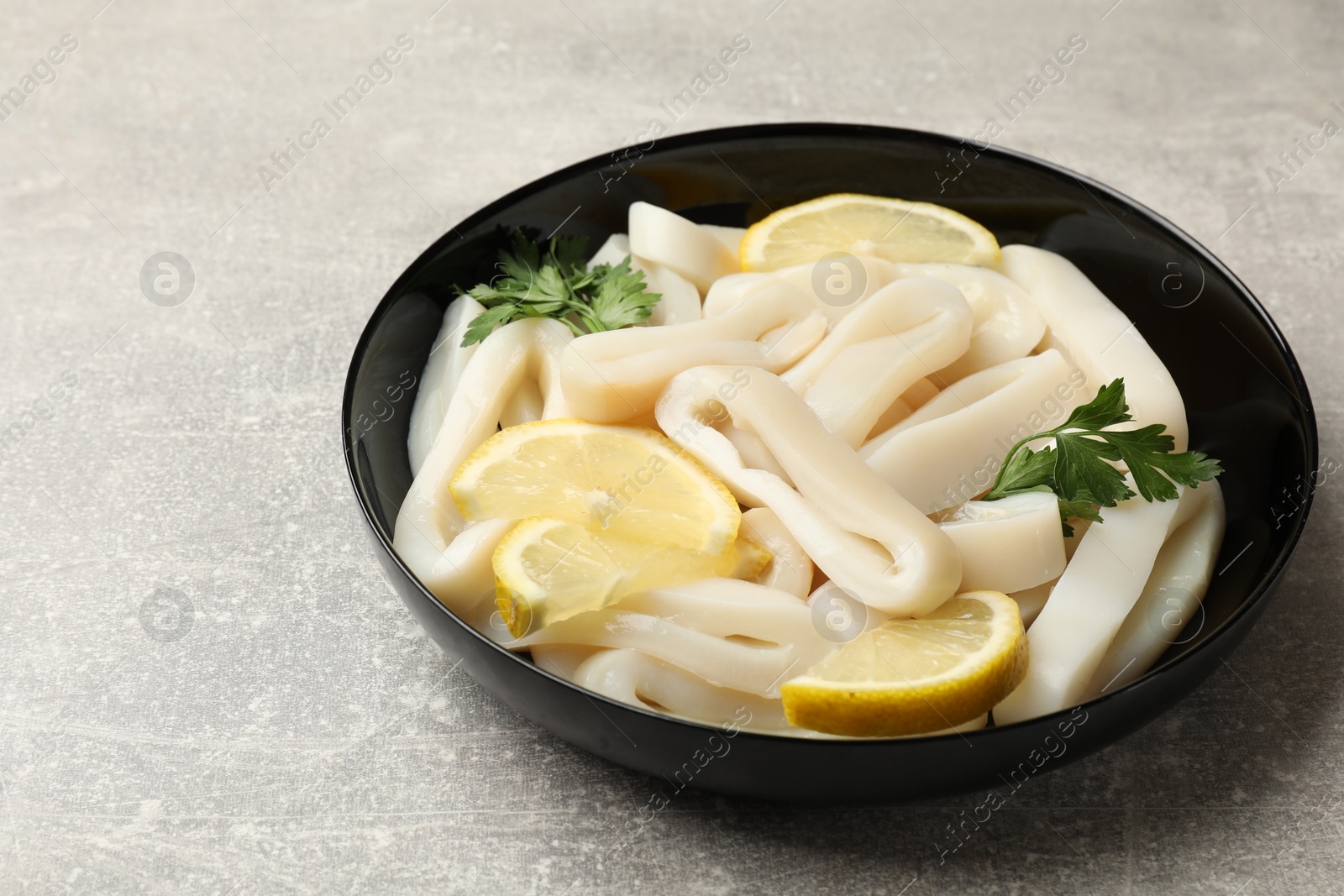 Photo of Raw squid rings, lemon and parsley in bowl on grey textured table, closeup. Space for text