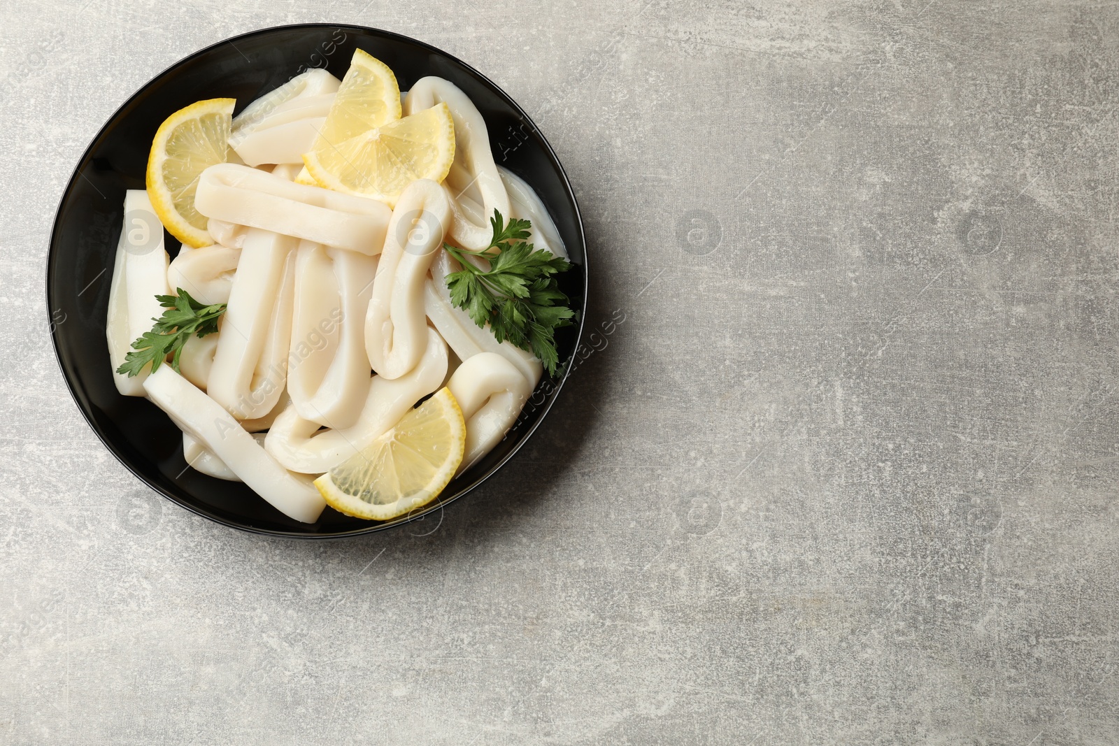 Photo of Raw squid rings, lemon and parsley in bowl on grey textured table, top view. Space for text