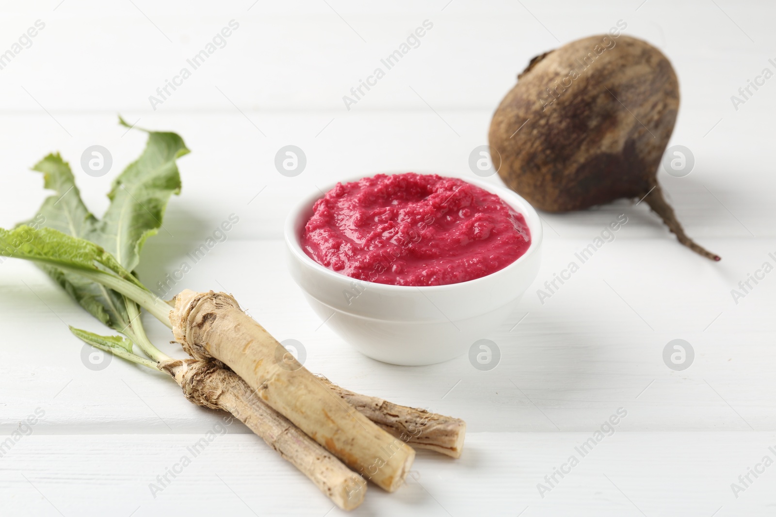Photo of Tasty horseradish sauce with beetroot, vegetable and roots on white wooden table, closeup