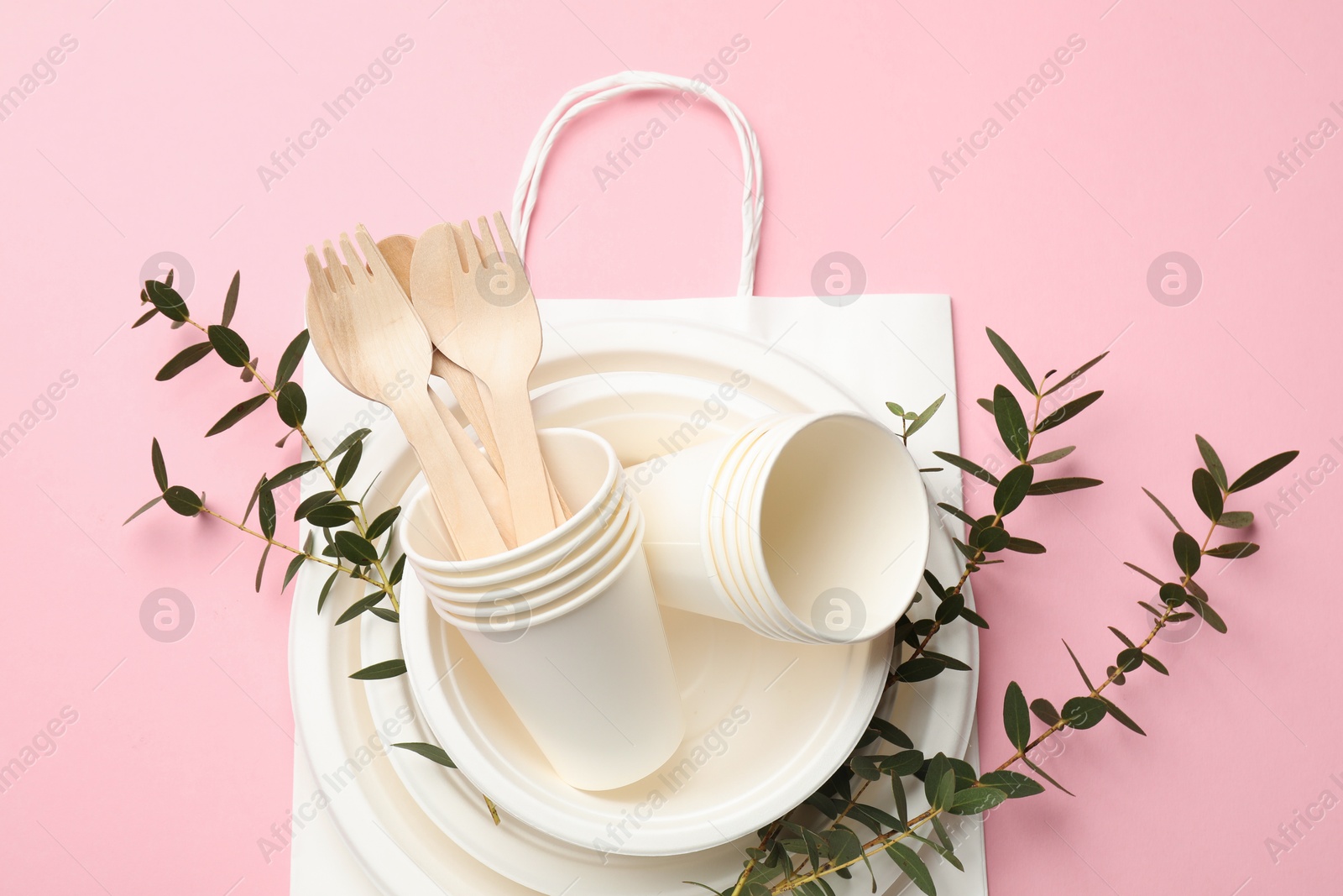 Photo of Set of disposable tableware, paper bag and eucalyptus branches on pink background, top view