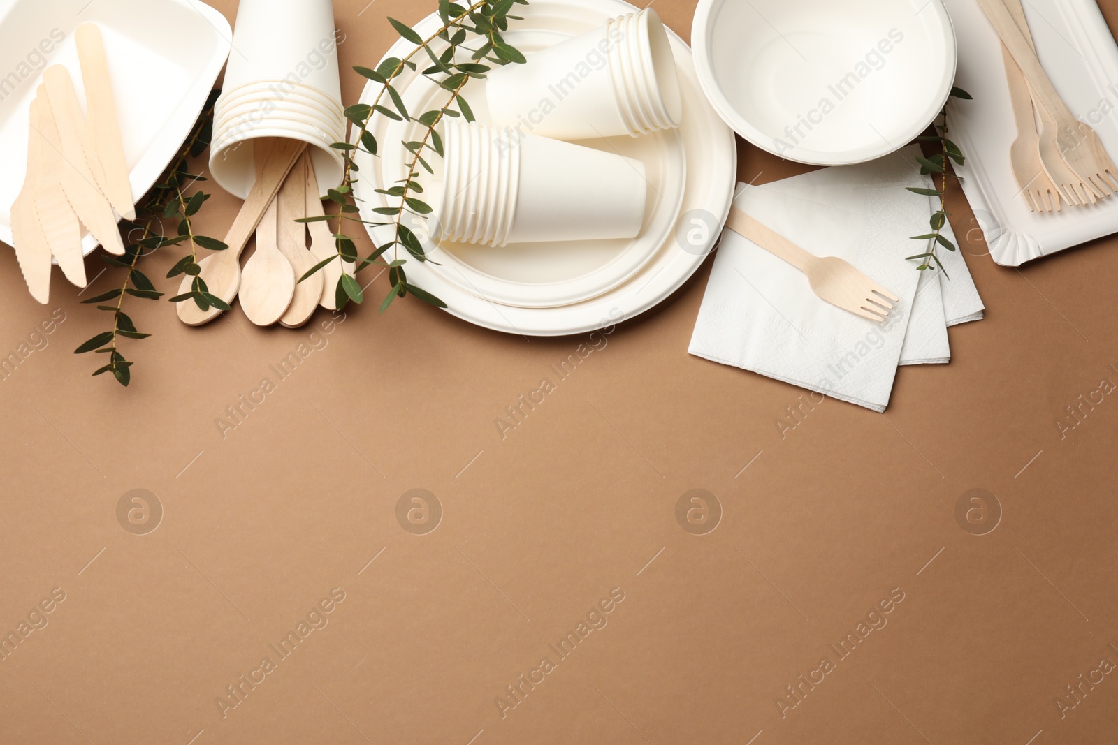 Photo of Set of disposable tableware and eucalyptus branches on brown background, flat lay. Space for text