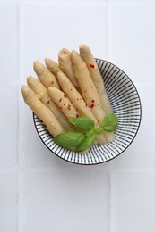 Photo of Pickled asparagus spears with spices and basil in bowl on white tiled table, top view