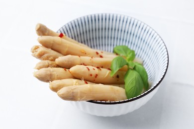 Photo of Pickled asparagus spears with spices and basil in bowl on white tiled table, closeup