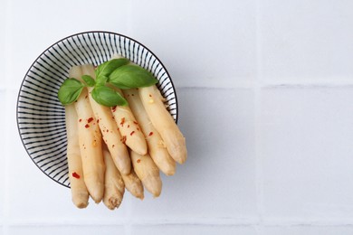 Photo of Pickled asparagus spears with spices and basil in bowl on white tiled table, top view. Space for text