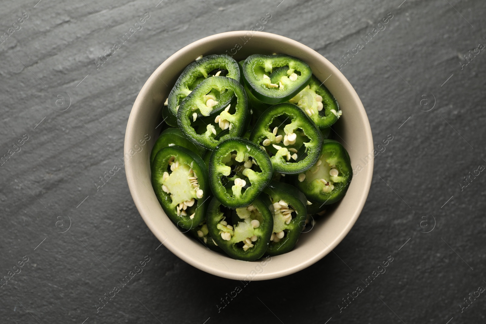Photo of Pieces of green jalapeno peppers in bowl on grey table, top view