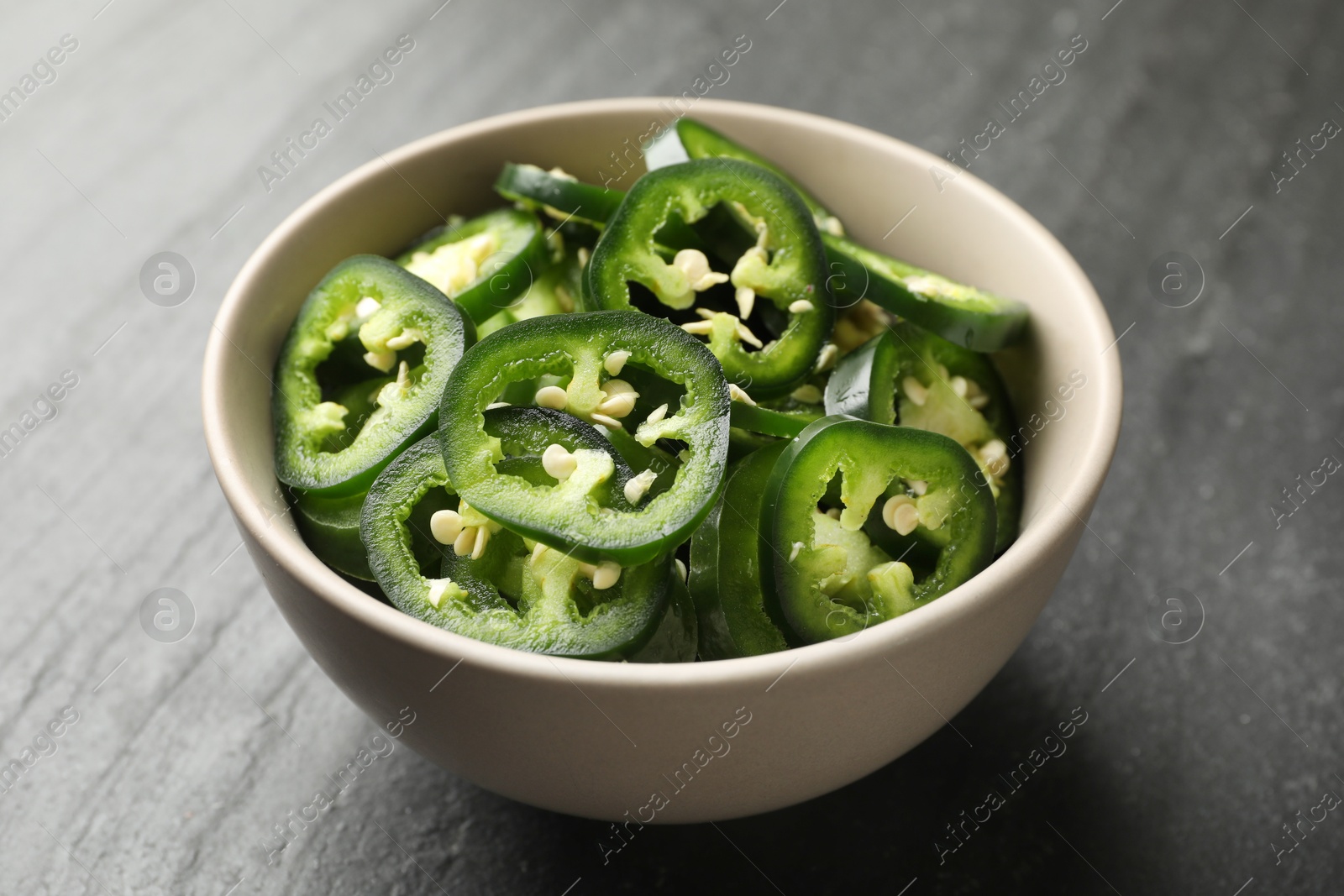 Photo of Pieces of green jalapeno peppers in bowl on grey table, closeup