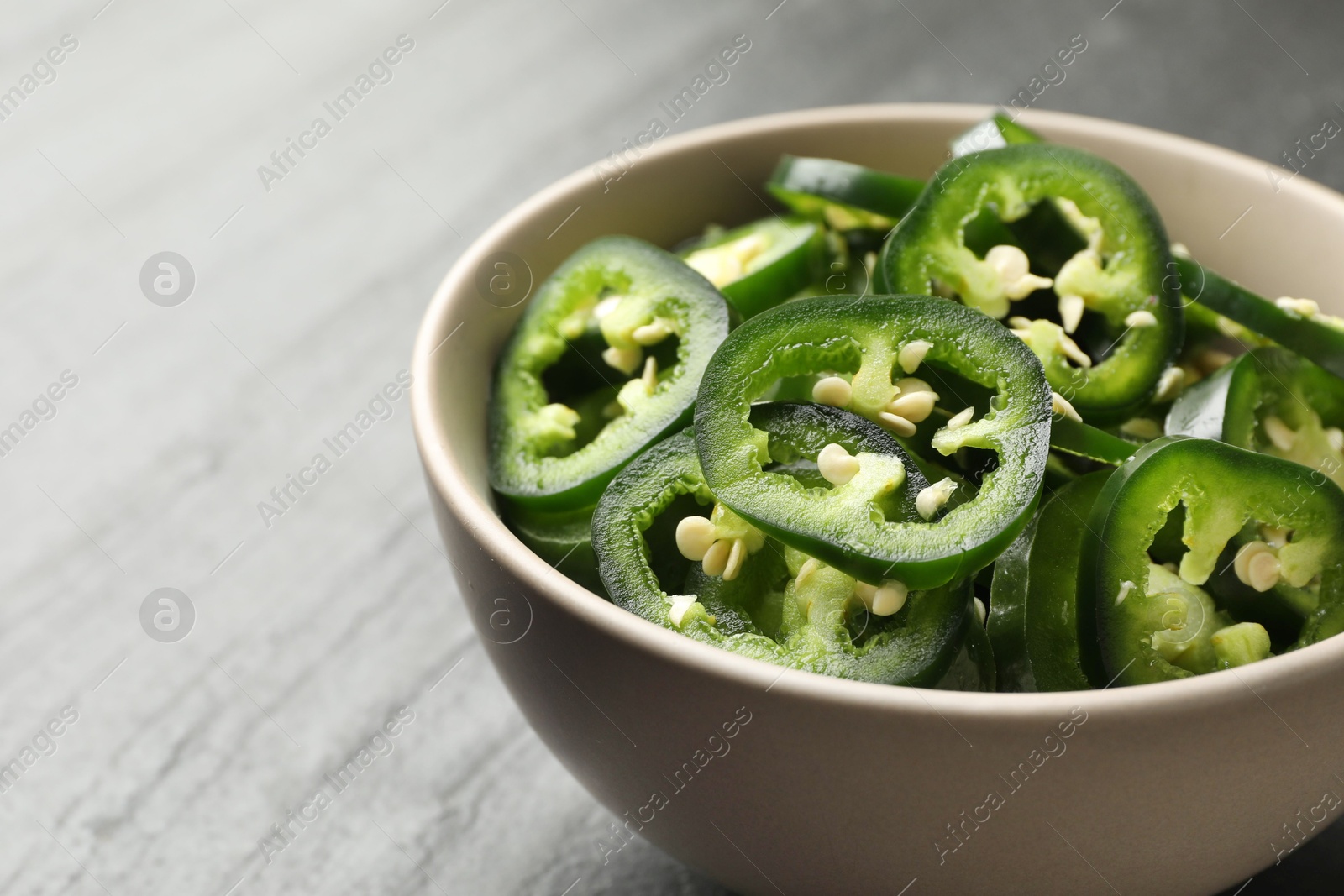 Photo of Pieces of green jalapeno peppers in bowl on grey table, closeup. Space for text