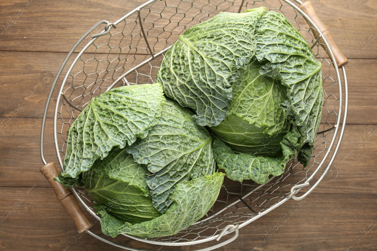 Photo of Fresh Savoy cabbages in metal basket on wooden table, top view