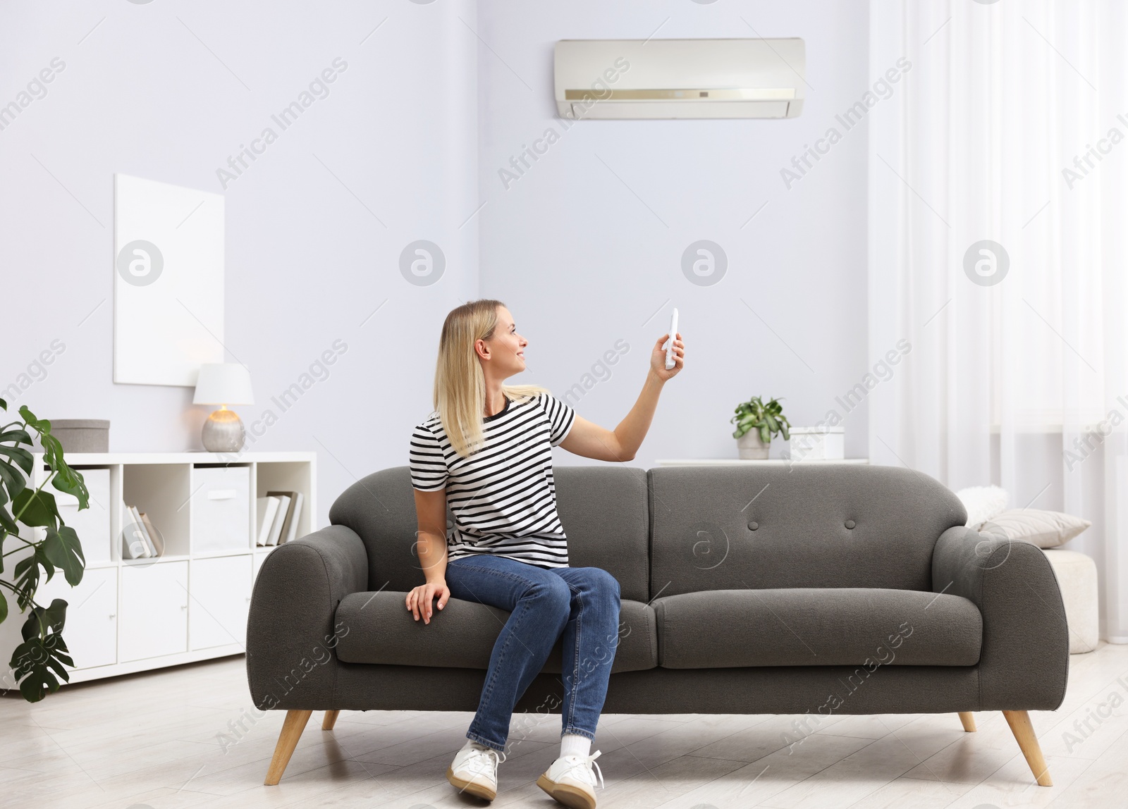 Woman using remote to control air conditioner on sofa at home Image of Woman using remote to control air conditioner on sofa at home