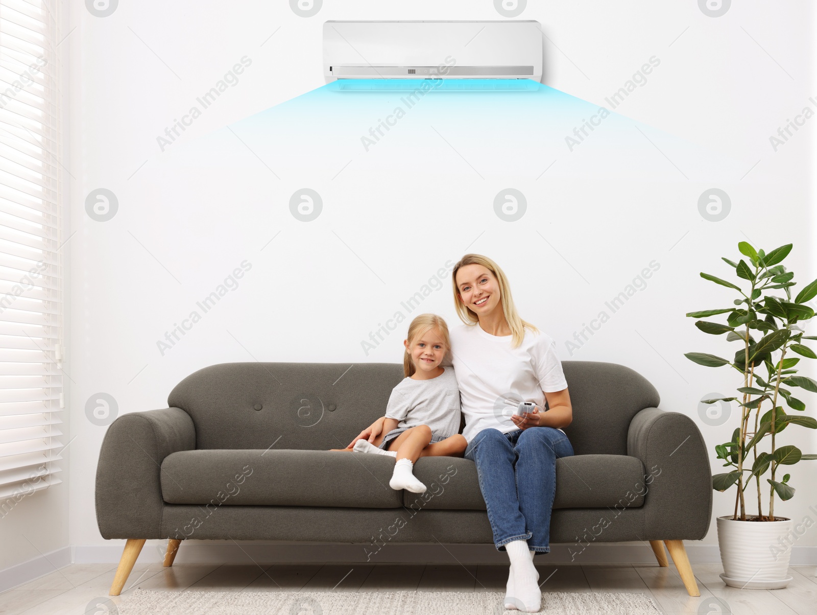 Image of Mother using remote to control air conditioner and hugging her daughter on sofa at home