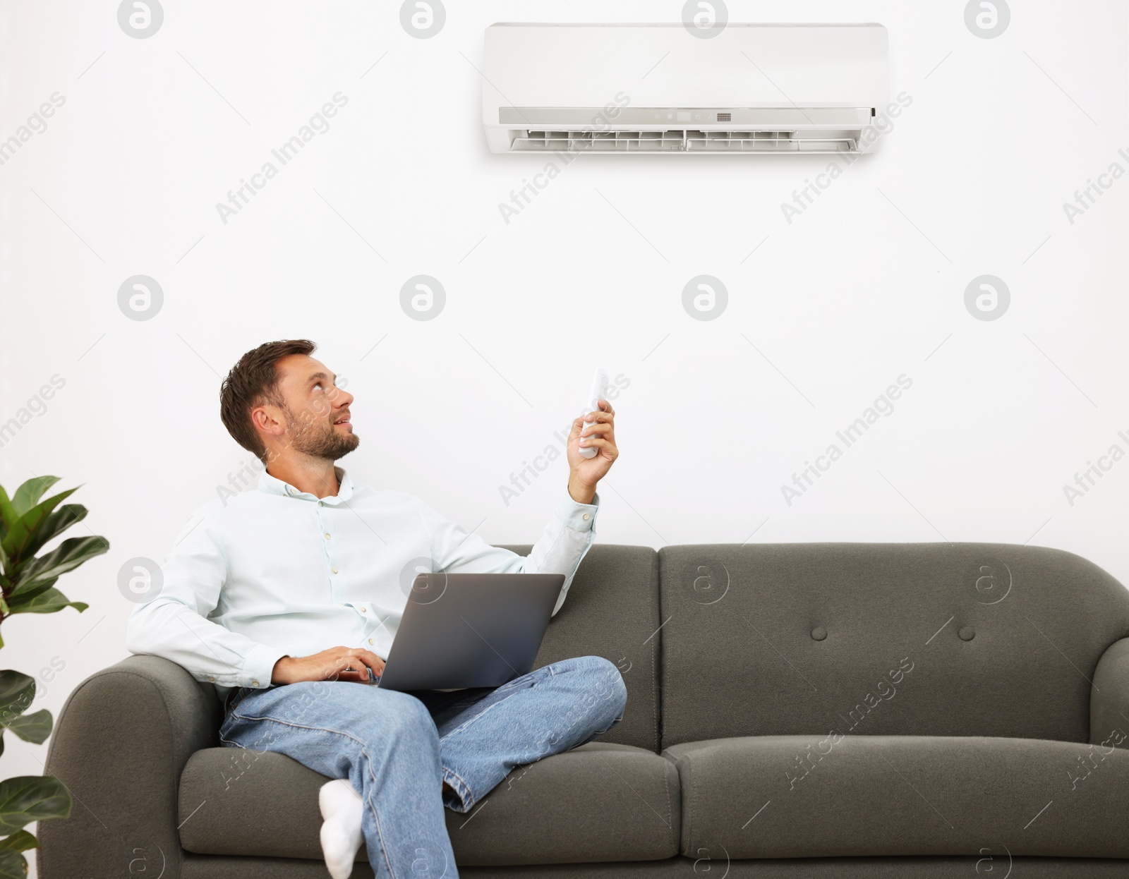 Image of Man with laptop using remote to control air conditioner on sofa at home
