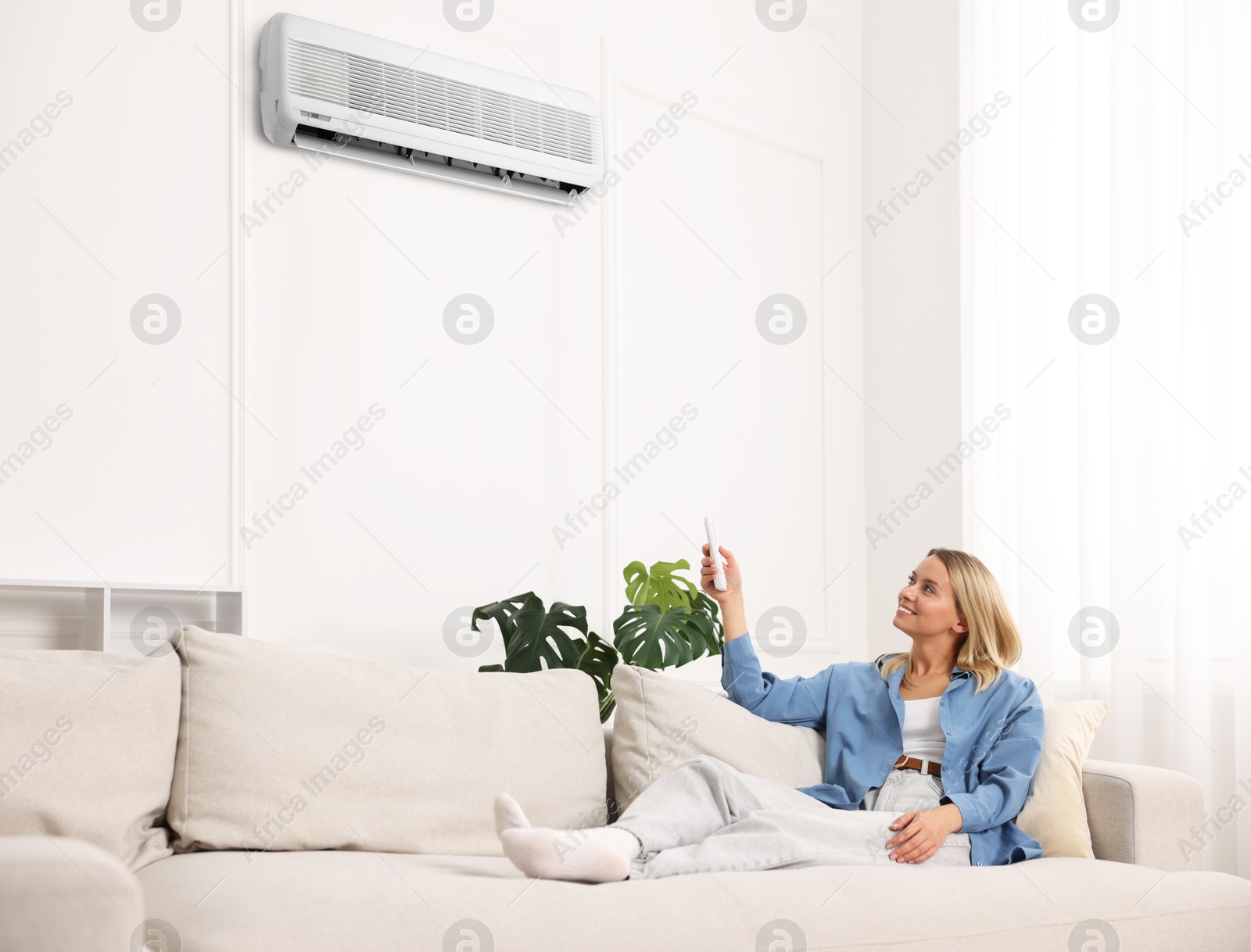 Image of Woman using remote to control air conditioner on sofa at home