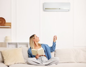 Image of Woman with book using remote to control air conditioner on sofa at home