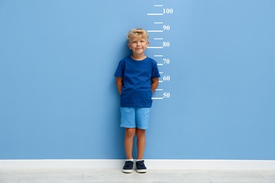 Image of Cute little boy measuring his height near blue wall with scale