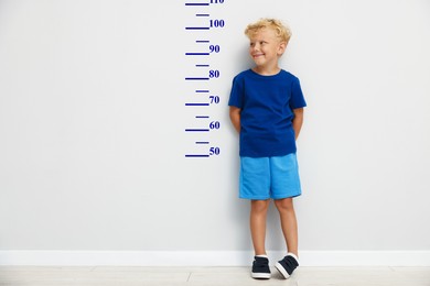 Image of Cute little boy measuring his height near white wall with scale