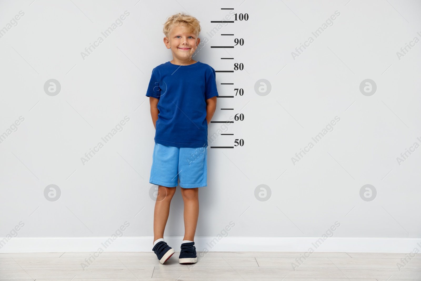 Image of Cute little boy measuring his height near white wall with scale