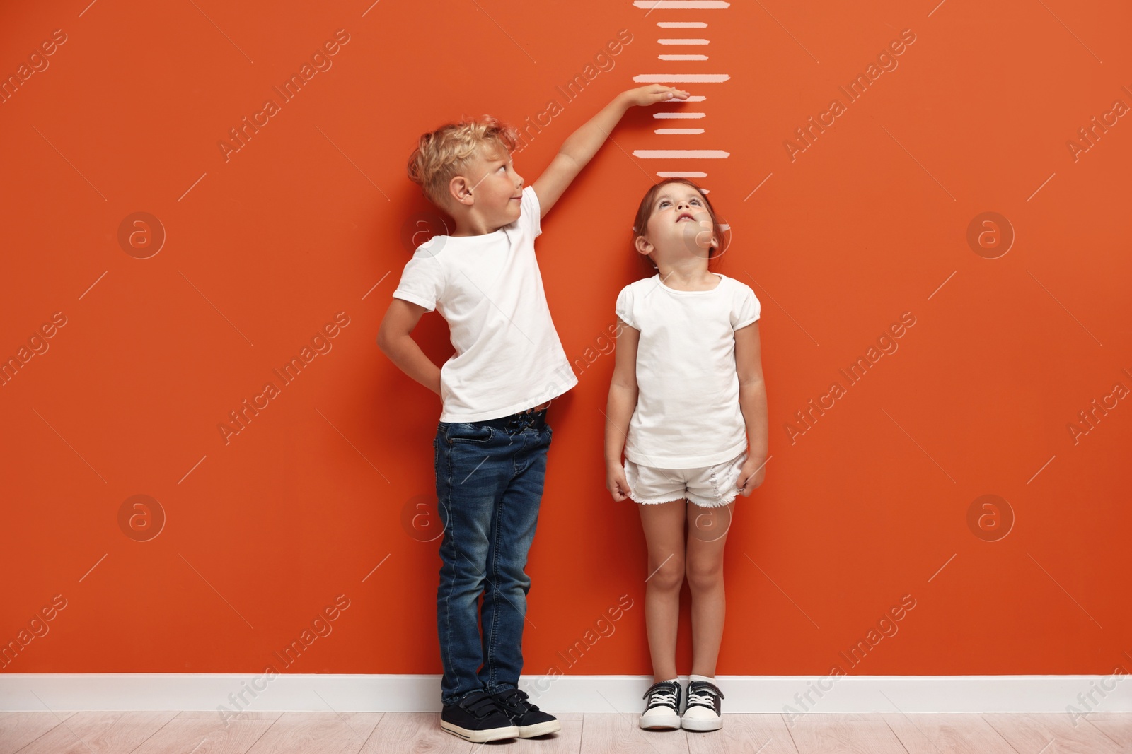 Image of Cute little girl and boy measuring their heights near orange wall with scale