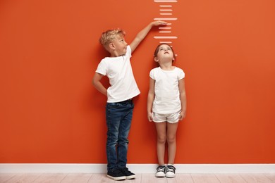 Image of Cute little girl and boy measuring their heights near orange wall with scale