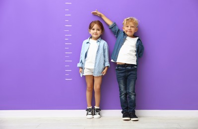 Image of Cute little girl and boy measuring their heights near violet wall with scale