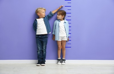 Image of Cute little girl and boy measuring their heights near violet wall with scale