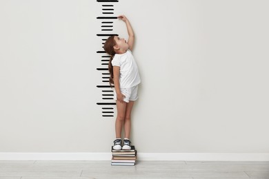 Image of Cute little girl on books measuring her height near white wall with scale