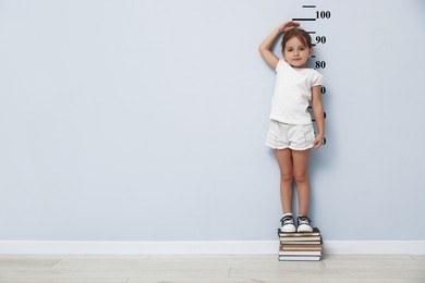 Image of Cute little girl on books measuring her height near white wall with scale