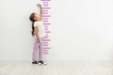 Image of Cute little girl measuring her height near white wall with scale