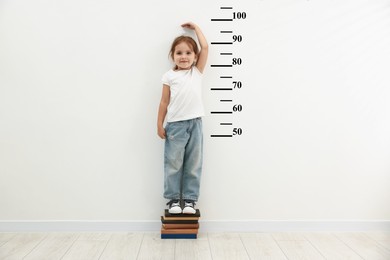 Image of Cute little girl on books measuring her height near white wall with scale