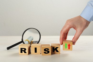 Image of Man putting wooden cube with arrows next to word Risk, magnifying glass and money on table