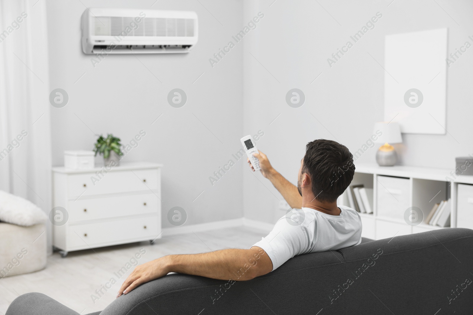 Image of Man using remote to control air conditioner on sofa at home, back view