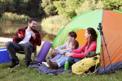 Photo of Happy family having break with camping tent, tourist equipment and drinks near river outdoors
