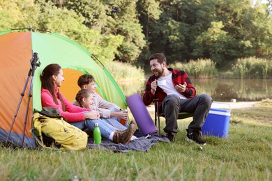Photo of Happy family having break with camping tent, tourist equipment and drinks near river outdoors